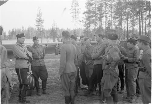 Vänrikki Mattila selostaa laivueen komentajalle, kapteeni Bremenille laivueen lentäjien ympäröimänä taisteluaan suuren venäläisen lentoveneen kanssa 14.7. Viim. rannikolla ryssä tuhoutui, Mattilan koneeseen 12 reikää